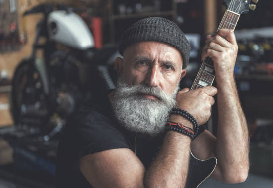 Confident bearded biker is siting in garage and holding black guitar. He glancing at camera with expectation. Portrait