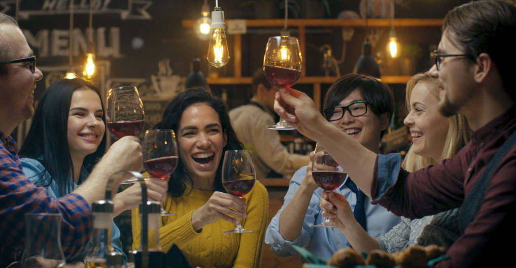 Diverse Group of Friends Celebrate with a Toast and Clink Raised Wine Glasses in Celebration. Beautiful Young People Have Fun in the Stylish Bar/ Restaurant.