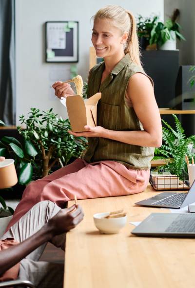 Young smiling female office manager eating wok and talking to African American male colleague sitting by workplace among green plants