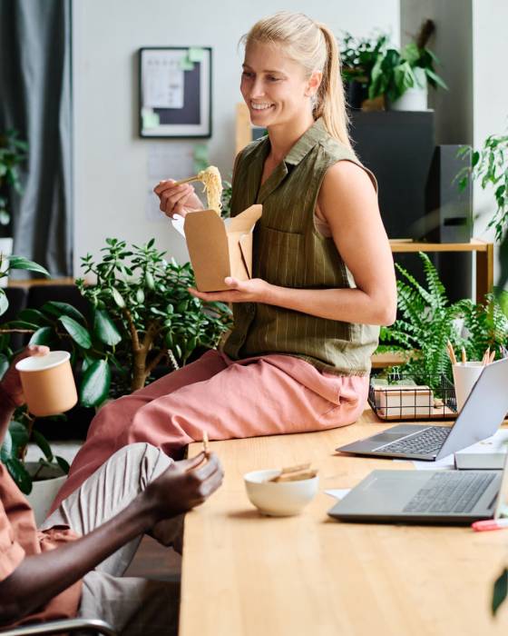 Young smiling female office manager eating wok and talking to African American male colleague sitting by workplace among green plants