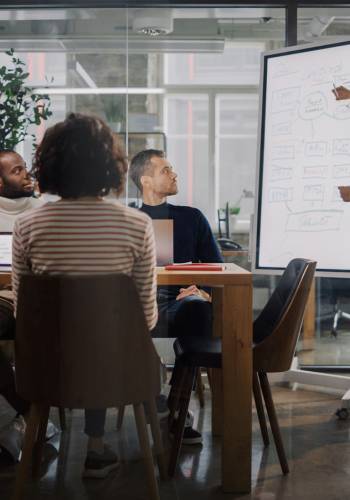 Project Manager Makes a Presentation for a Young Diverse Creative Team in Meeting Room in an Agency. Colleagues Sit Behind Conference Table and Discuss Business Development, User Interface and Design.