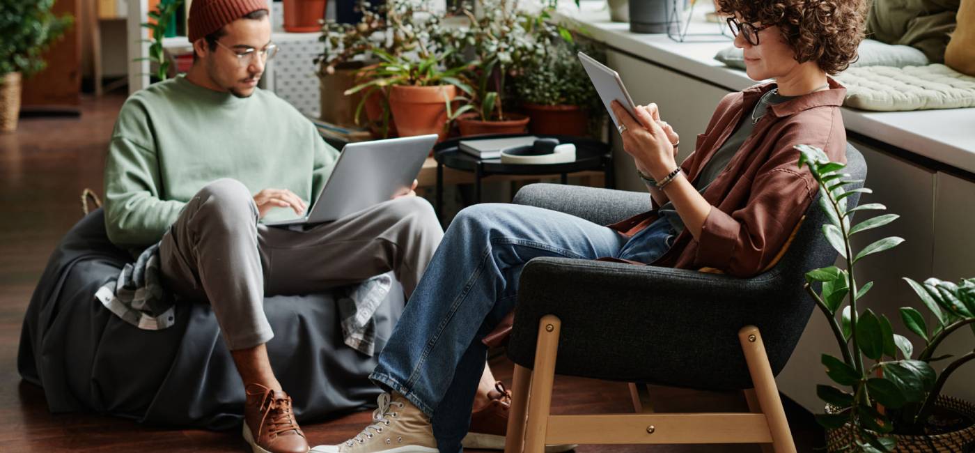 Two young serious coworkers using wireless mobile gadgets while sitting in armchairs in openspace office with variety og green plants