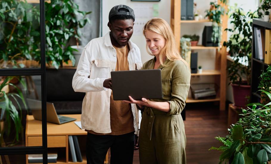 Two young intercultural employees in casualwear looking at screen of laptop held by blond businesswoman during discussion