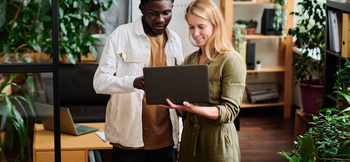 Two young intercultural employees in casualwear looking at screen of laptop held by blond businesswoman during discussion