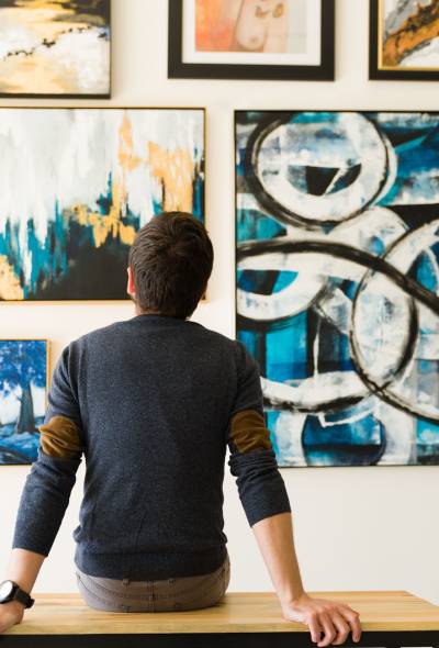 Hispanic male visitor looking reflective while sitting on a bench and admiring the various paintings on the wall of an art gallery