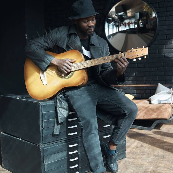 Musician in black trench coat and bucket hat sitting on bed and playing guitar. Songwriter in loft room. Fashion 2019