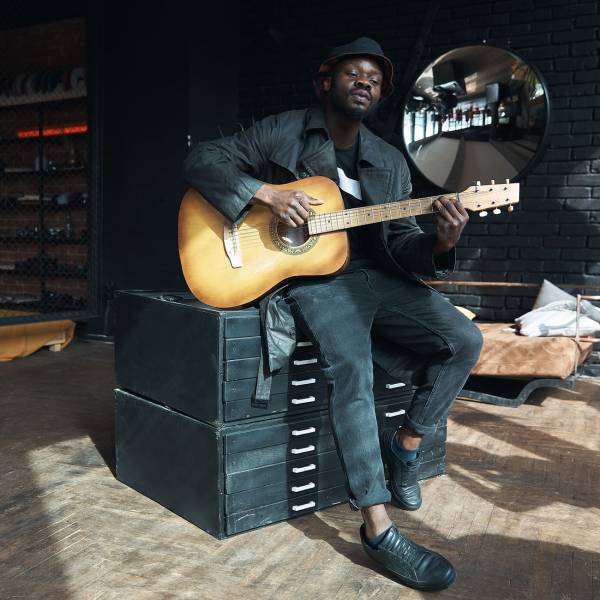 Musician in black trench coat and bucket hat sitting on bed and playing guitar. Songwriter in loft room. Fashion 2019