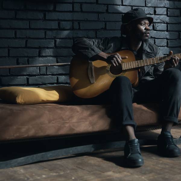 Musician in black trench coat and bucket hat sitting on bed and playing guitar. Songwriter in loft room. Fashion 2019