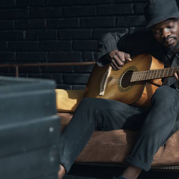 Musician in black trench coat and bucket hat sitting on bed and playing guitar. Songwriter in loft room. Fashion 2019