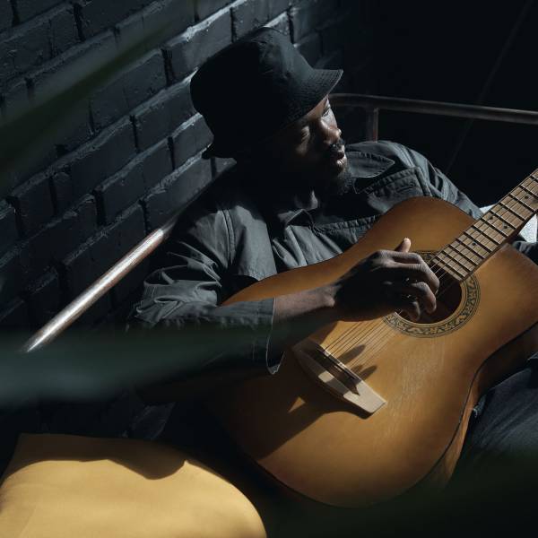 Musician in black trench coat and bucket hat sitting on bed and playing guitar. Songwriter in loft room. Fashion 2019