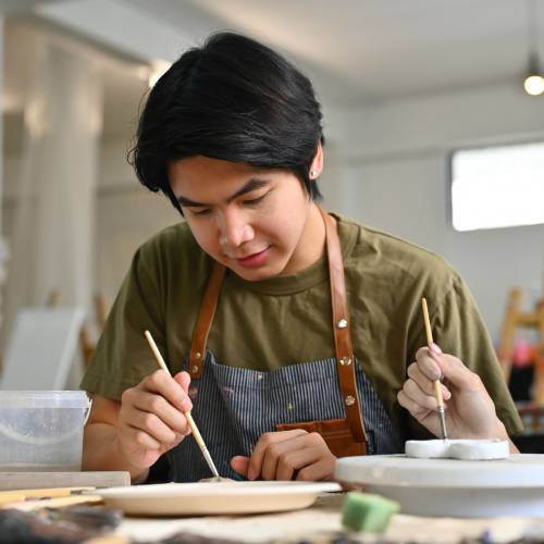 Two teenage Asian students focused on painting ceramic pieces in an art classroom together.